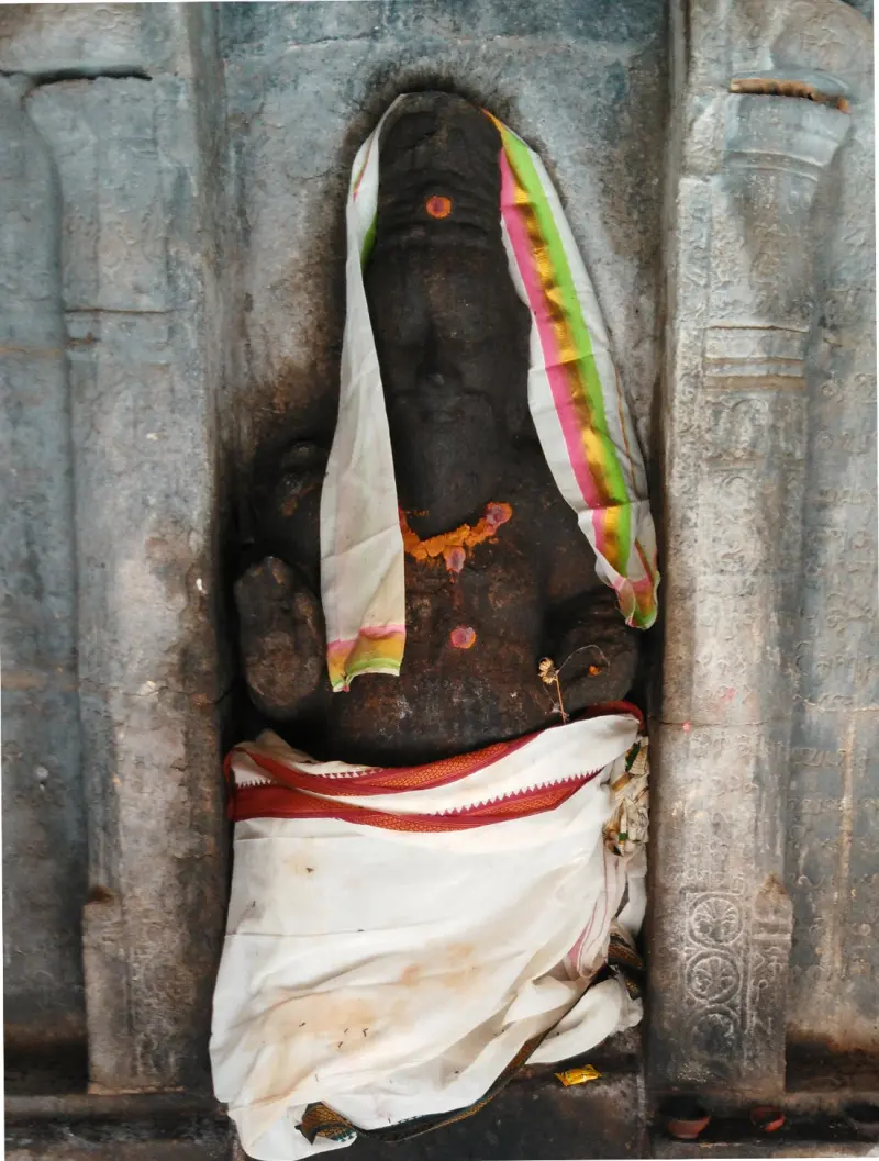 A dark stone Vinayakar draped in a white cloth and a striped shawl sits in a carved niche.