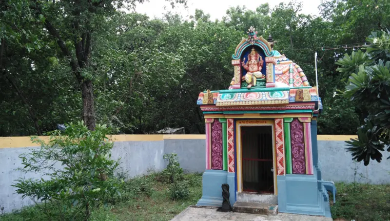 A small painted shrine under trees, with a seated Vinayakar figure above its doorway