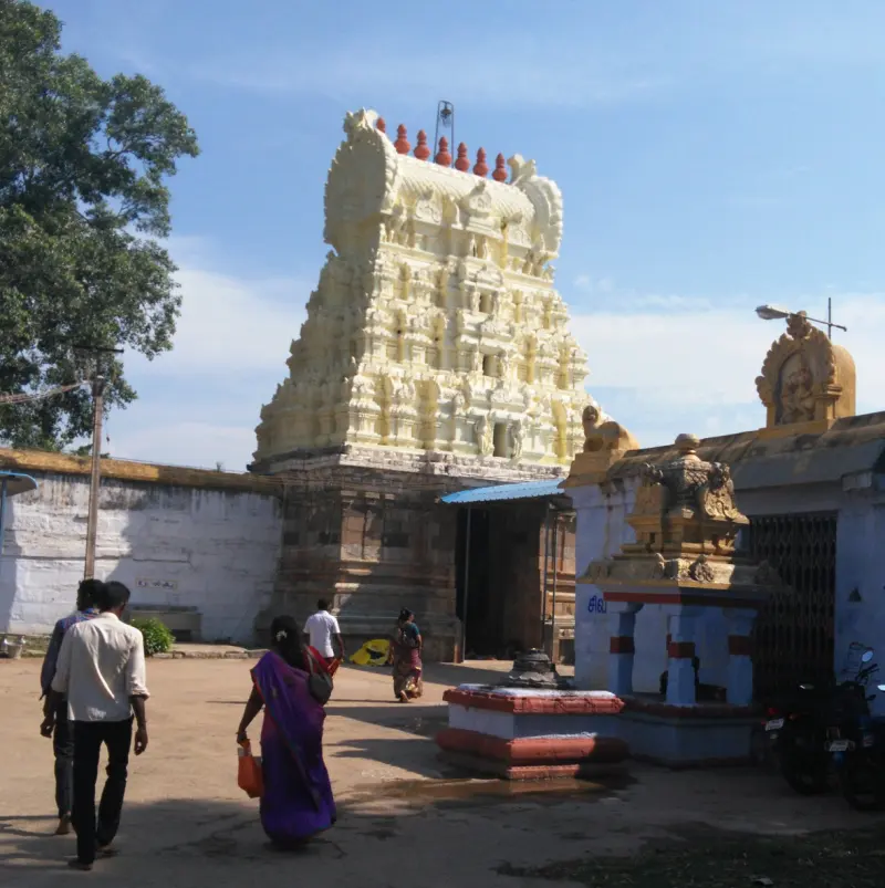 A cream-coloured tiered gopuram over a stone base, with devotees walking in the courtyard.