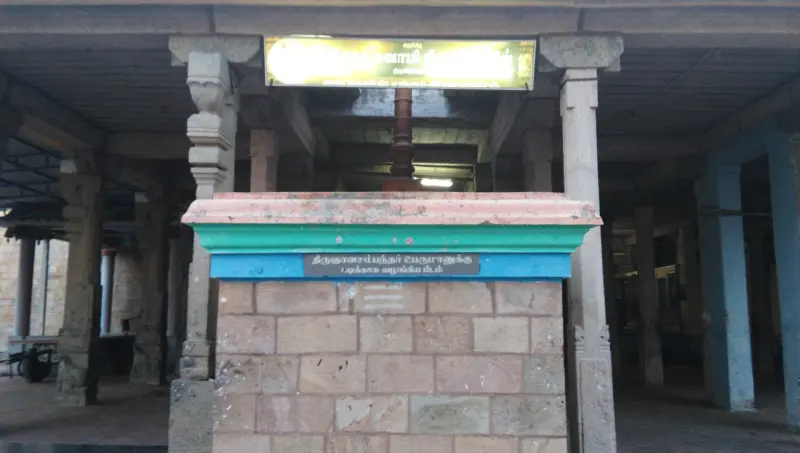 A stone block with a coloured top and a Tamil name plate stands in a pillared hall; a flagstaff base is behind it.