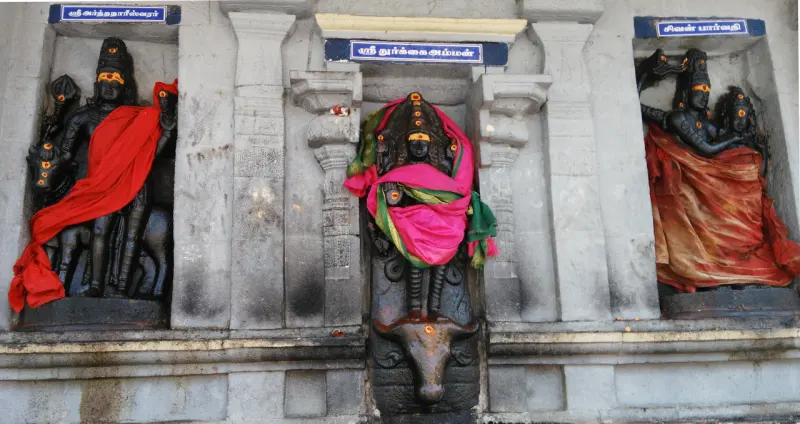 Three carved figures on a grey wall: Ardhanareeswarar, Durgai above a bull's head, and Shiva with Parvathi