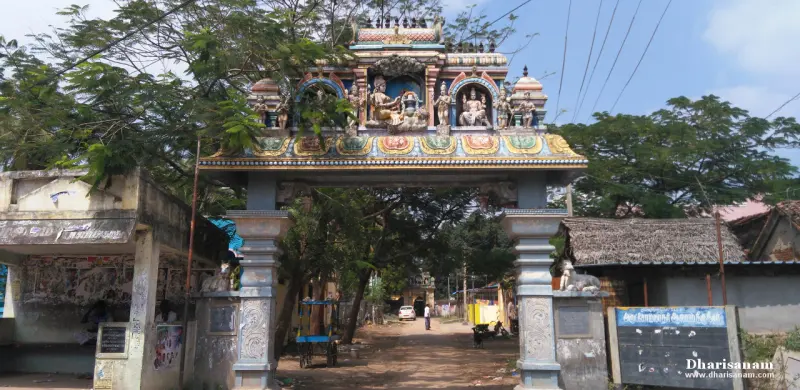 A painted gateway arch with seated deity sculptures under a tiled roof, over a village road lined with trees