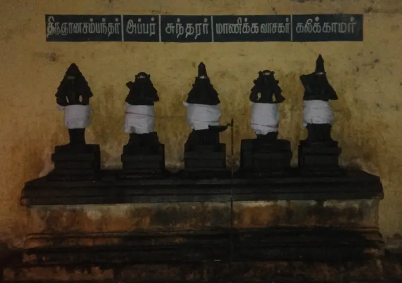 Five small dark figures in white cloth on a stone ledge, with a row of Tamil names above