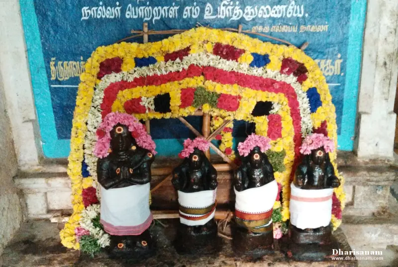 Four small dark idols in white cloths and flower garlands standing before a flower arch