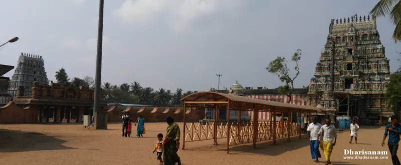 A wide courtyard with a covered walkway, one tower at left and a colourful one at right, devotees walking