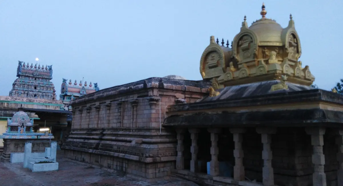 A golden domed tower above a stone sanctum with a pillared hall in front; a gopuram rises at the far left at dusk.