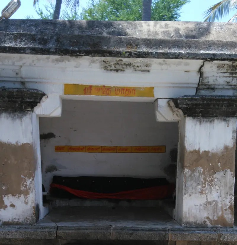 A whitewashed open shrine with a yellow Tamil name board and a dark image lying under a red cloth.