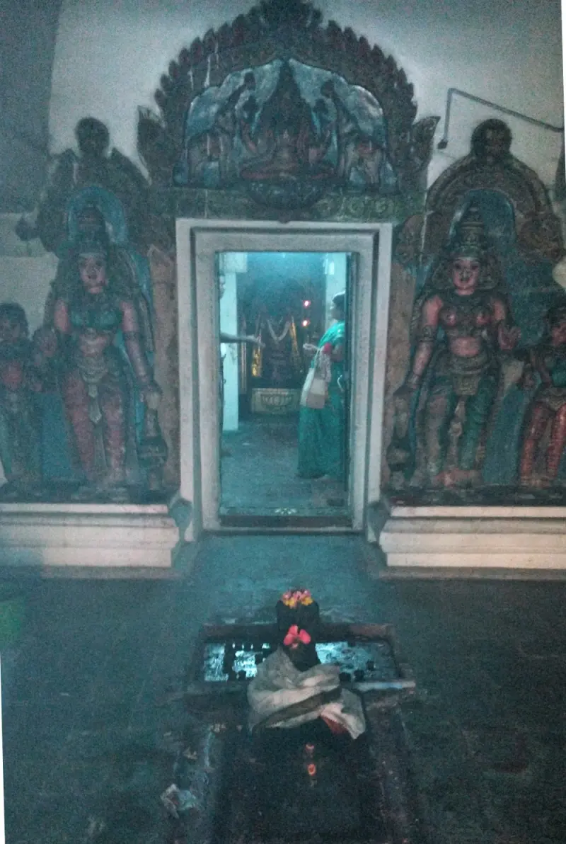 A doorway shows a woman inside a dim hall; painted figures flank it, and a small garlanded stone lies in front.