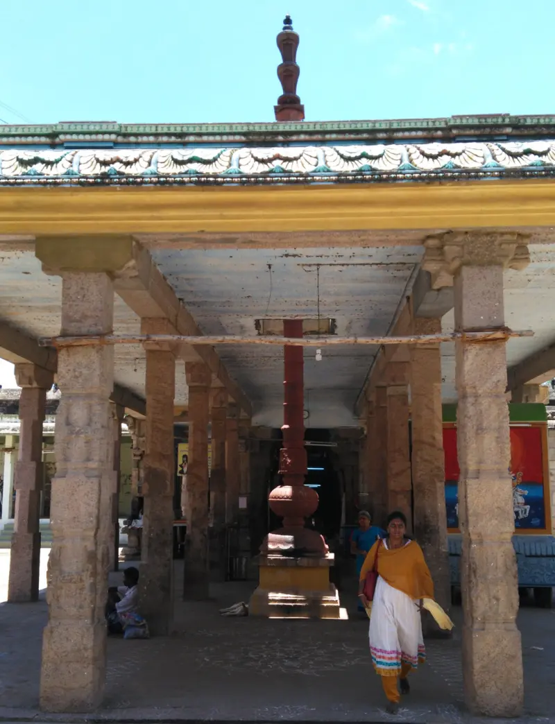 A stone pillared hall with a tall red flagstaff on a base at the centre and a few people walking through.