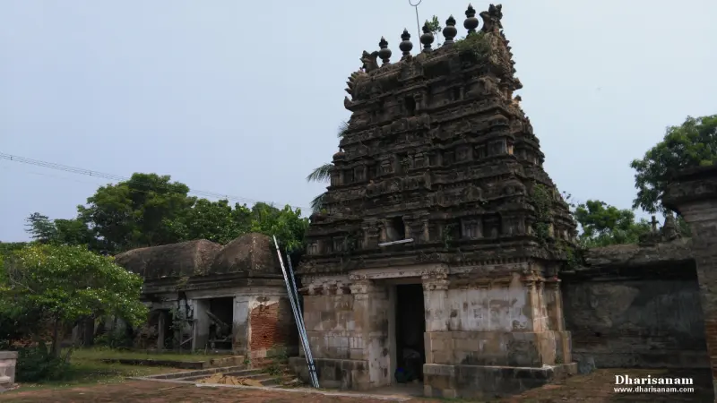 A dark, weathered gopuram with finials and plants growing on its tiers, beside a ruined hall at left.