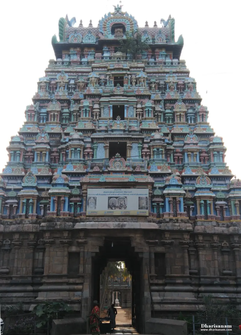 A tiered gopuram above a stone gateway, with two people walking in the passage beyond