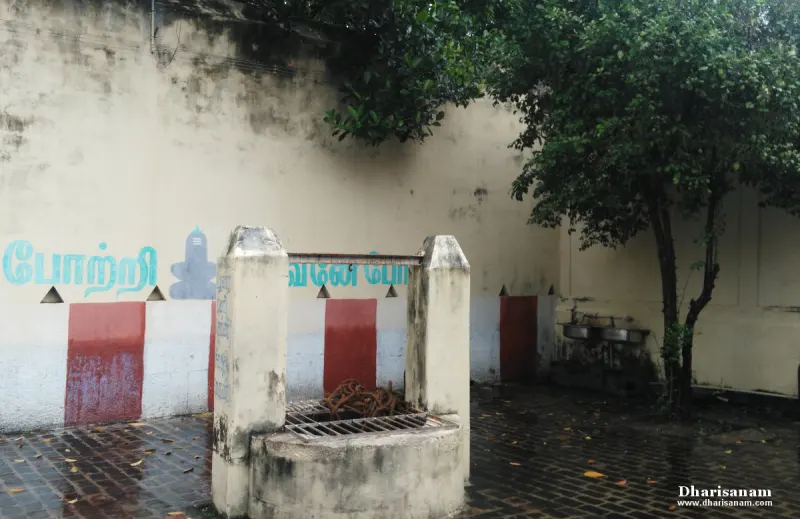 Stone well with a metal grille beside a whitewashed wall with a Tamil inscription and a tree