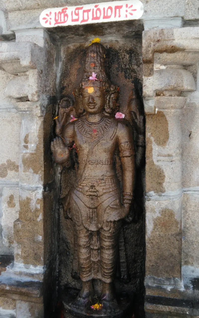 A standing stone image of Brahma in a niche, adorned with flowers, under a Tamil name board