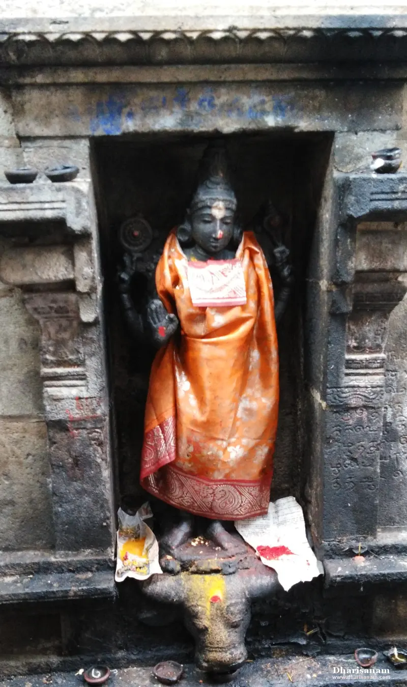 A dark stone idol in an orange silk cloth stands in a niche above a carved animal head, with lamps around