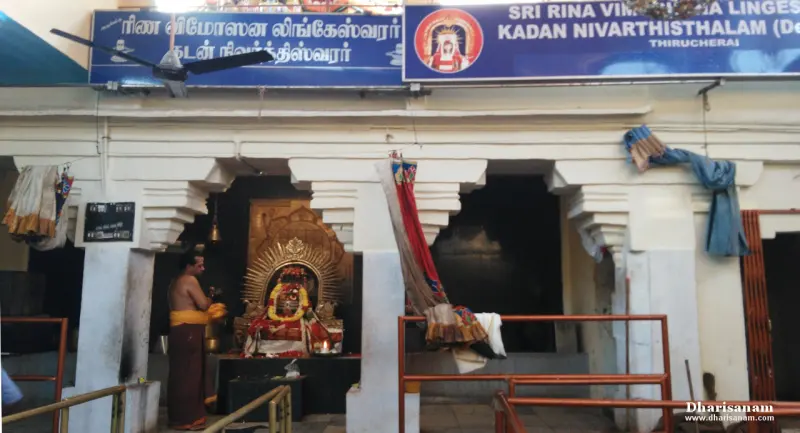 A decorated Shivalingam in a pillared shrine, a priest beside it and a signboard above naming the lord