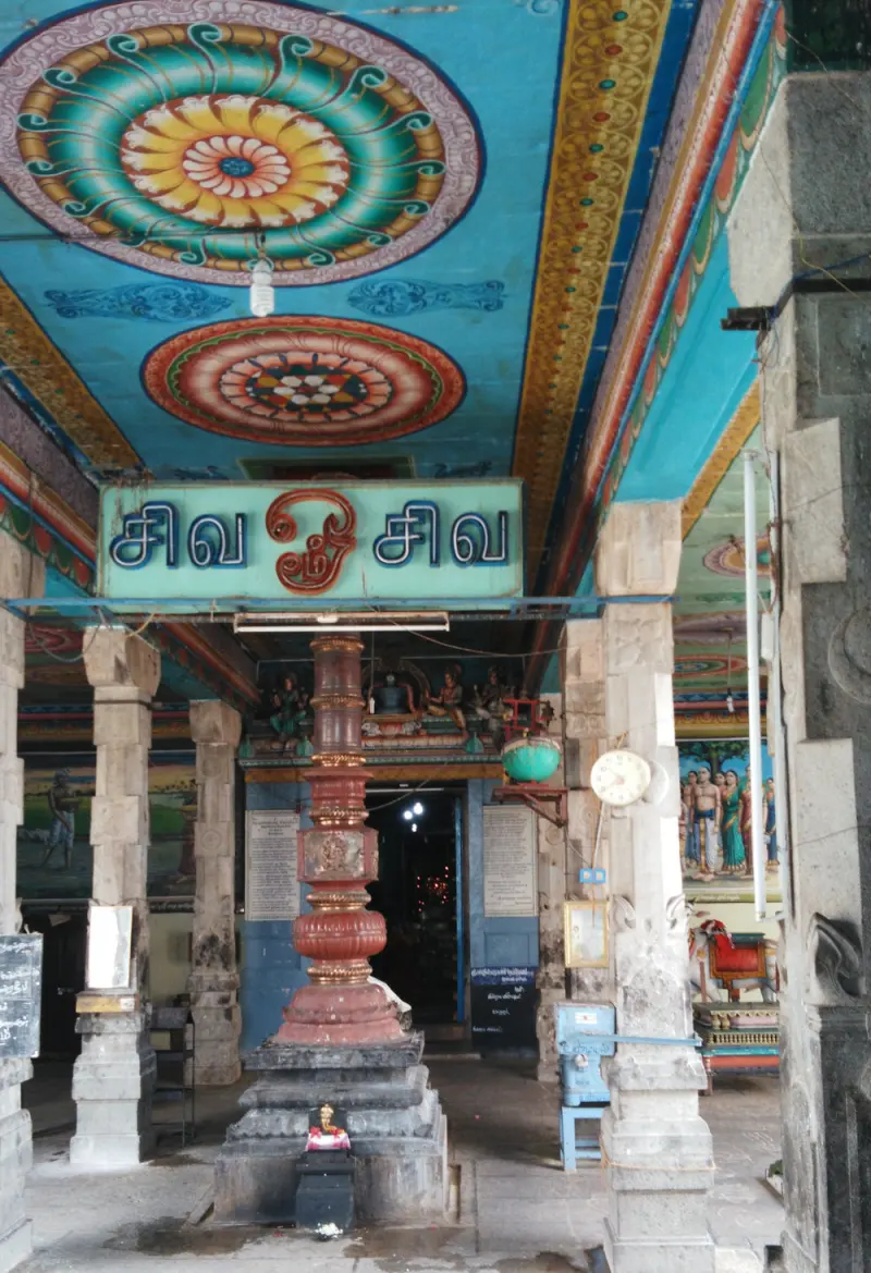 A flagstaff base in a hall with a brightly painted ceiling and a Tamil sign above the passage.
