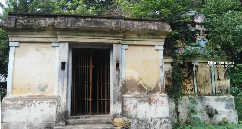 A small shrine with plain walls and a closed iron gate; a painted tower and figure show above the wall at right.