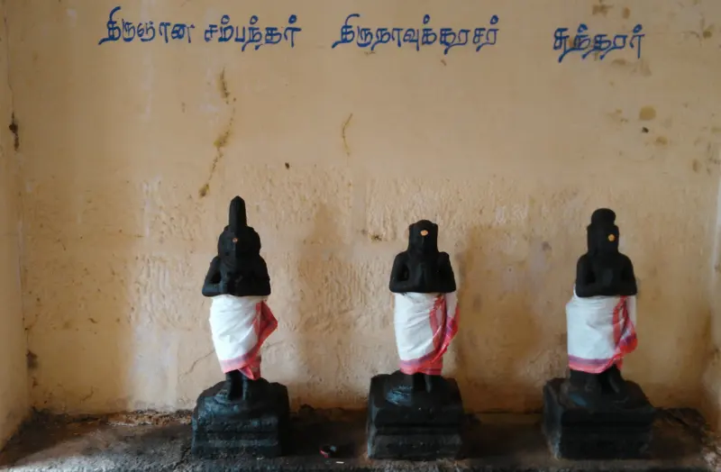 Three small black idols in white cloth stand in a row beneath Tamil name labels
