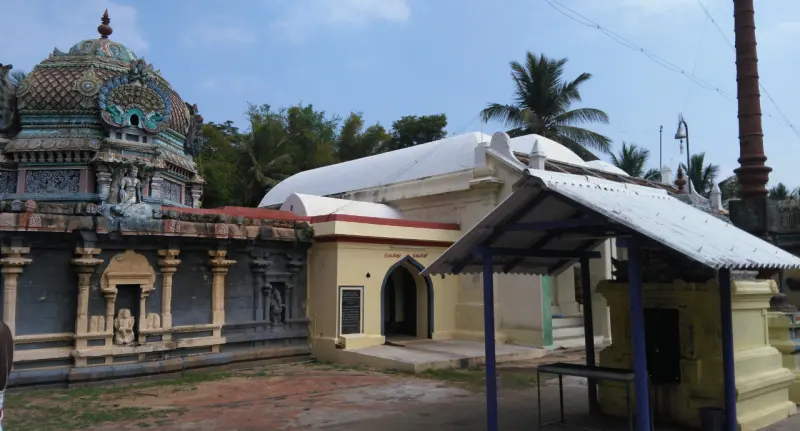 A yellow shrine under a tin roof stands in a paved courtyard, beside a long carved stone wall and a tall flagstaff.