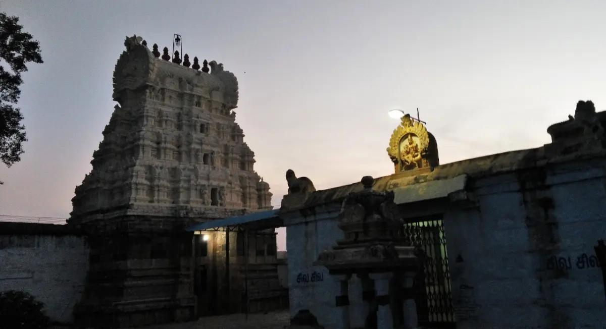 A tiered stone gopuram beside a long temple wall, with a lit golden arch on the roof, at dusk.