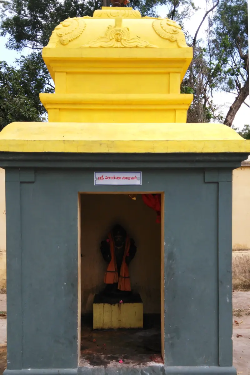 A small grey shrine with a yellow roof and a name plate; a dark idol in an orange cloth stands inside