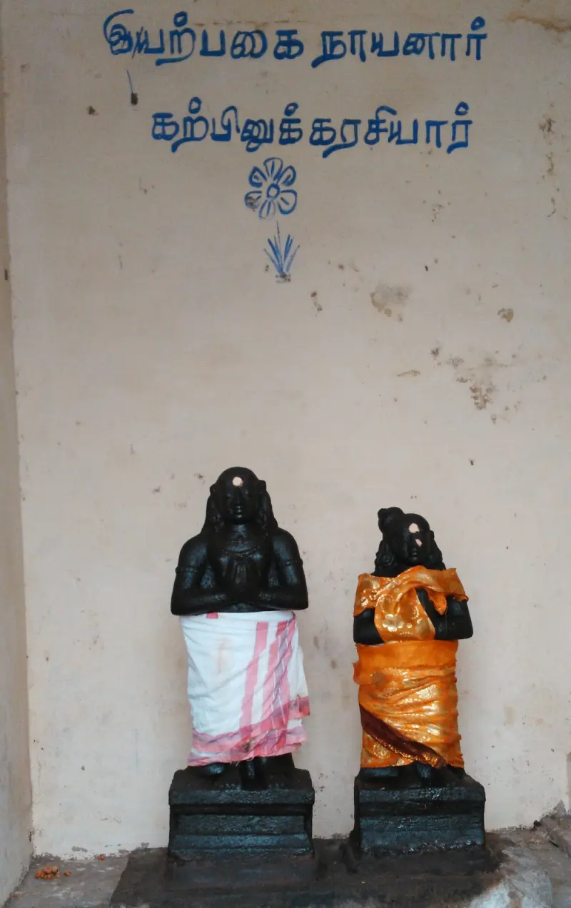 Two small idols stand before a wall labelled Eyarpagai Nayanar and Karpinukarasiyar
