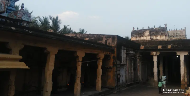 A covered walkway of stone pillars, with a person walking and an old tower top in the distance.