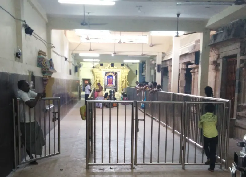 A long hall with steel barriers across the floor; devotees stand near a lit golden shrine at the far end.