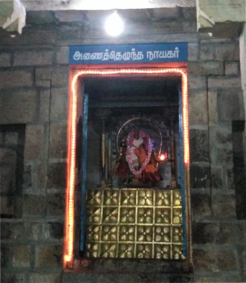 A lit niche in a stone wall holds a red-garlanded idol, under a blue Tamil board naming the deity