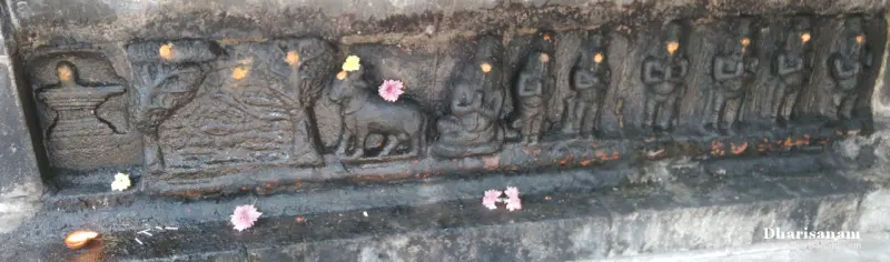A long stone relief with a lingam, a tree, a cow and rows of small seated and standing figures, with fresh flowers