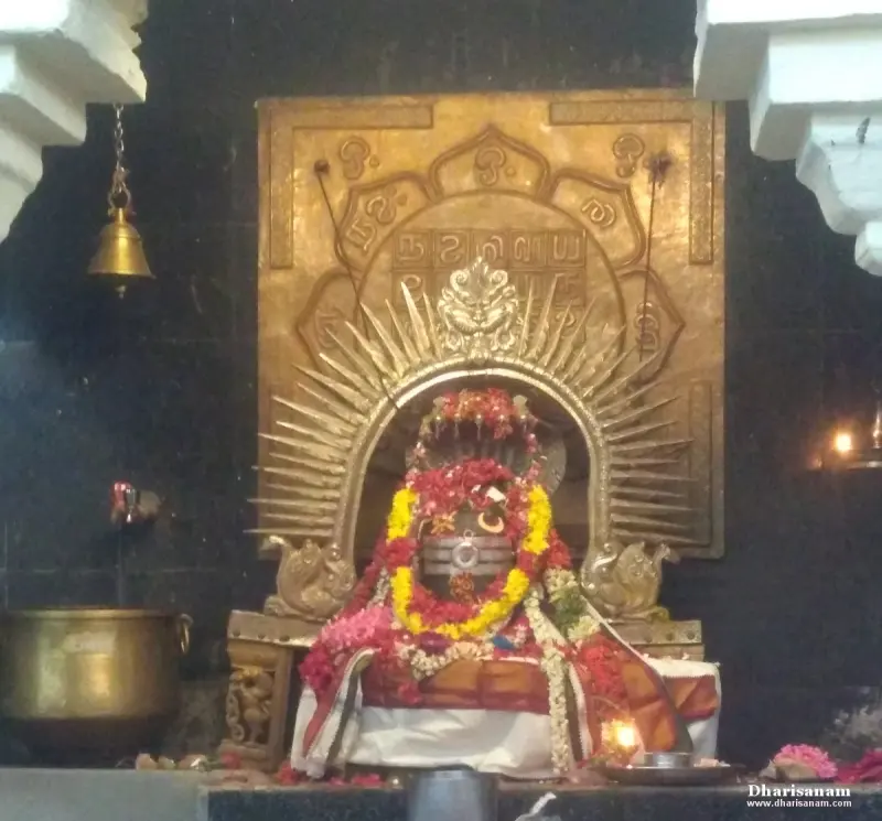 A garlanded Shivalingam under a golden arch with a bell and a large brass vessel beside it