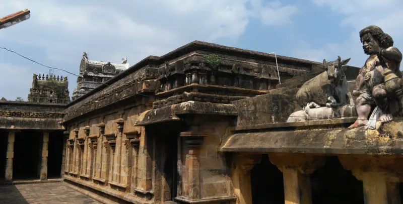 A stone Nandi and a seated figure on the ledge of a pillared corridor; gateway towers in the distance.