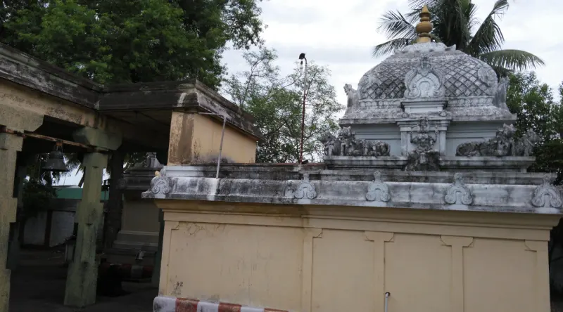 A small shrine with a carved stone dome and a golden finial, seen above a yellow wall, with trees behind.