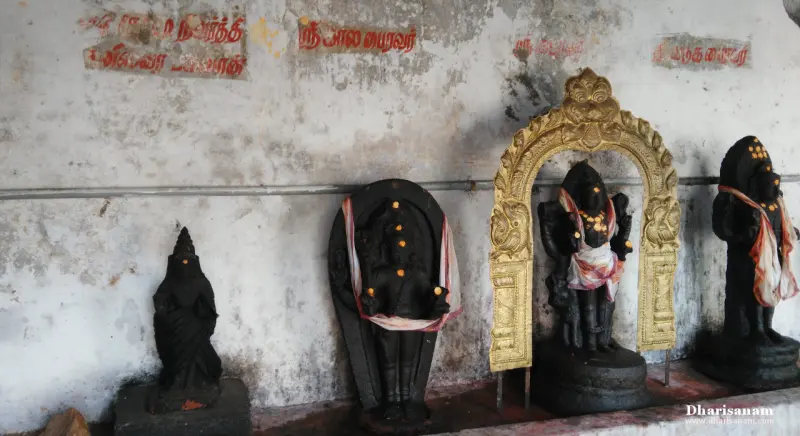 Dark stone idols in a row below Tamil labels on a weathered wall; one stands inside a gilded arch