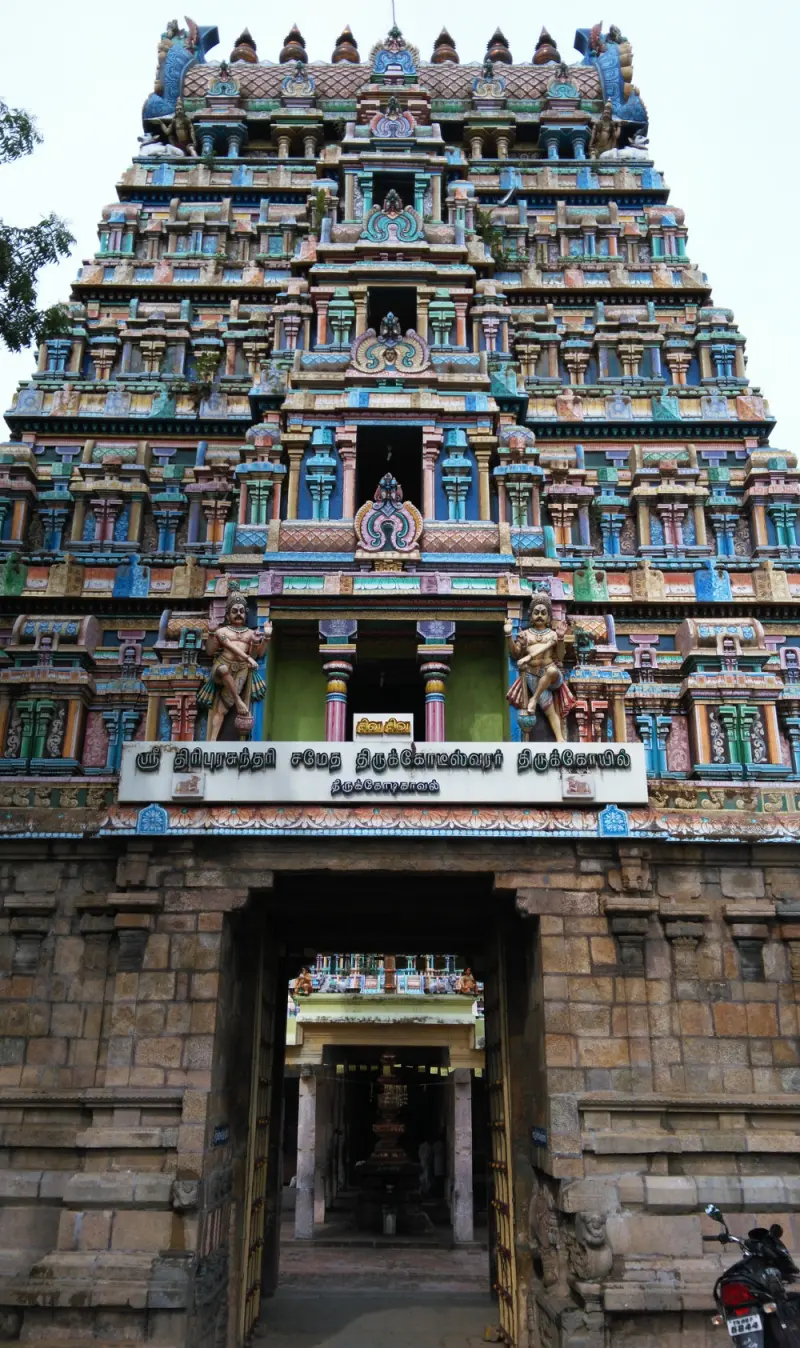 A tall, colourful tower above a stone gateway, with a Tamil name board and guardian figures at its base.