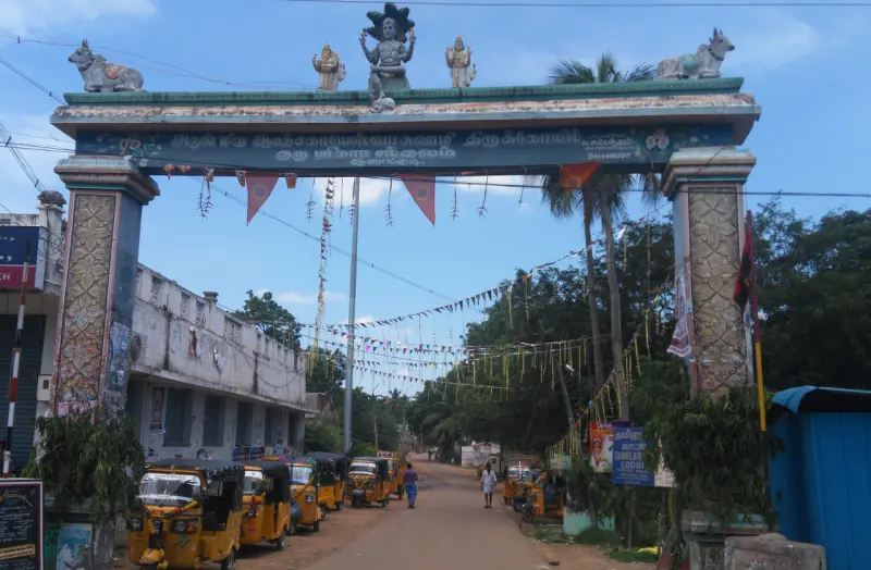 A tall road arch with a Tamil name board and figures on top, over a street with auto-rickshaws