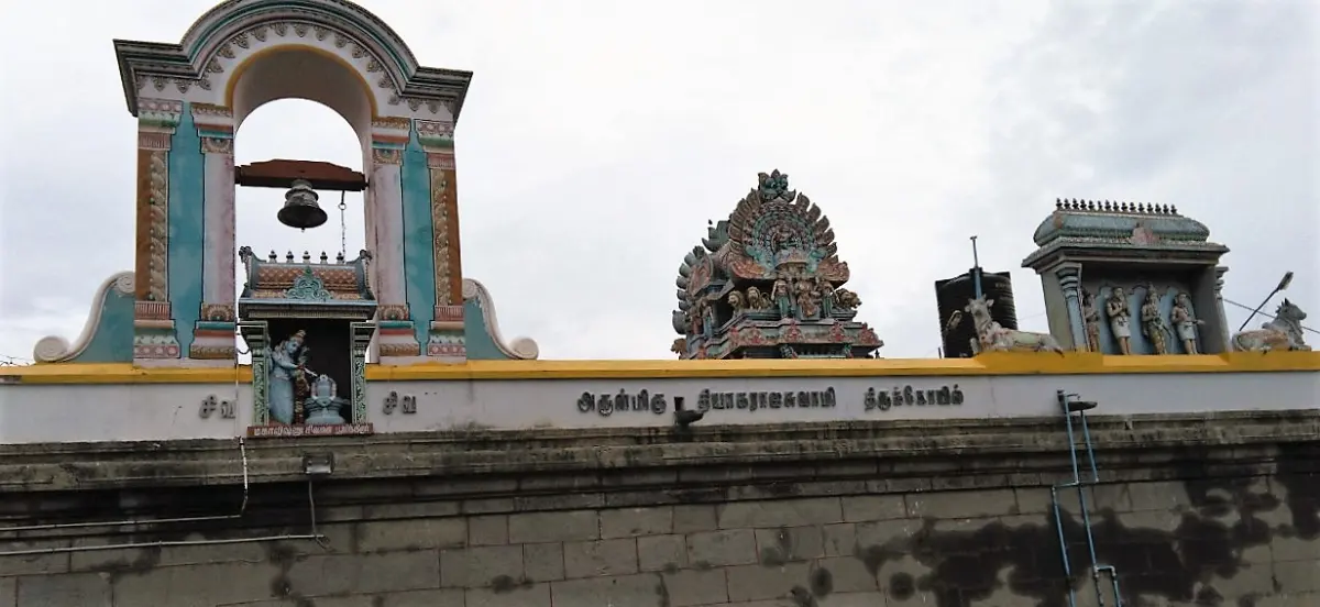A long wall with a bell hanging in an arch, a small shrine below it and painted towers behind.