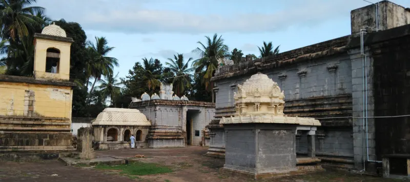 Small domed shrines in an open courtyard, a yellow building with a tower at the left and palms behind.