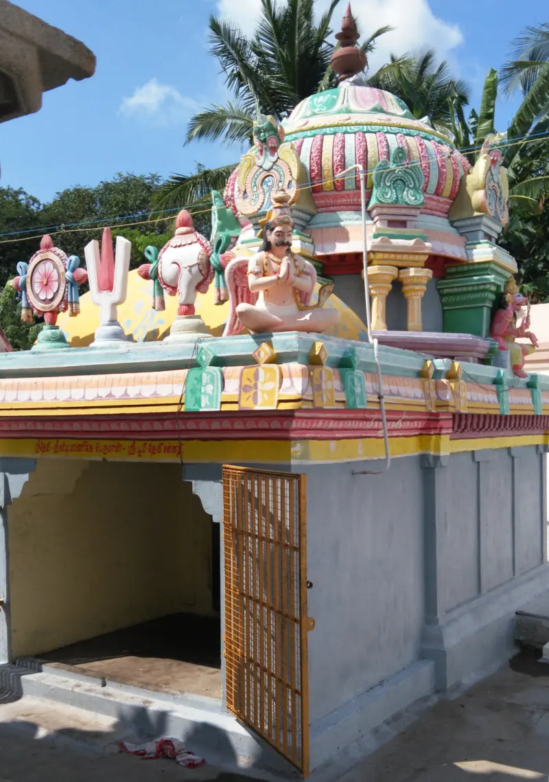 A shrine whose roof carries a seated stucco figure with folded hands, with its door grille open