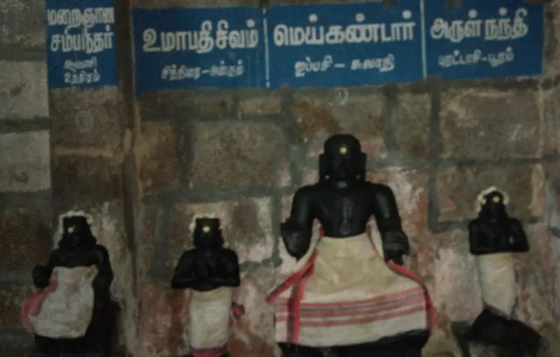 Four black idols in white cloth stand in a row beneath blue Tamil boards naming each saint