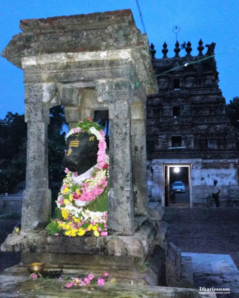 A dark stone Nandi decorated with flower garlands in a pillared pavilion; the gopuram and its gateway stand behind.