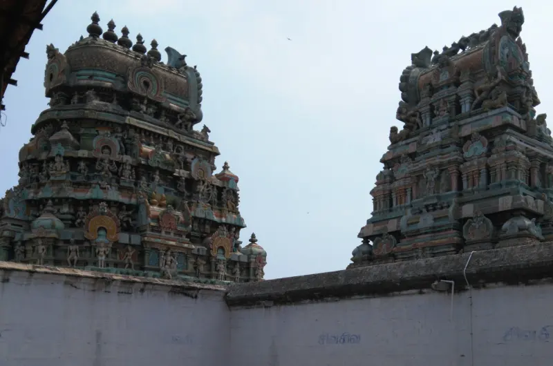 Two colourful towers covered in painted figures rise above a plain white wall with Tamil writing on it.