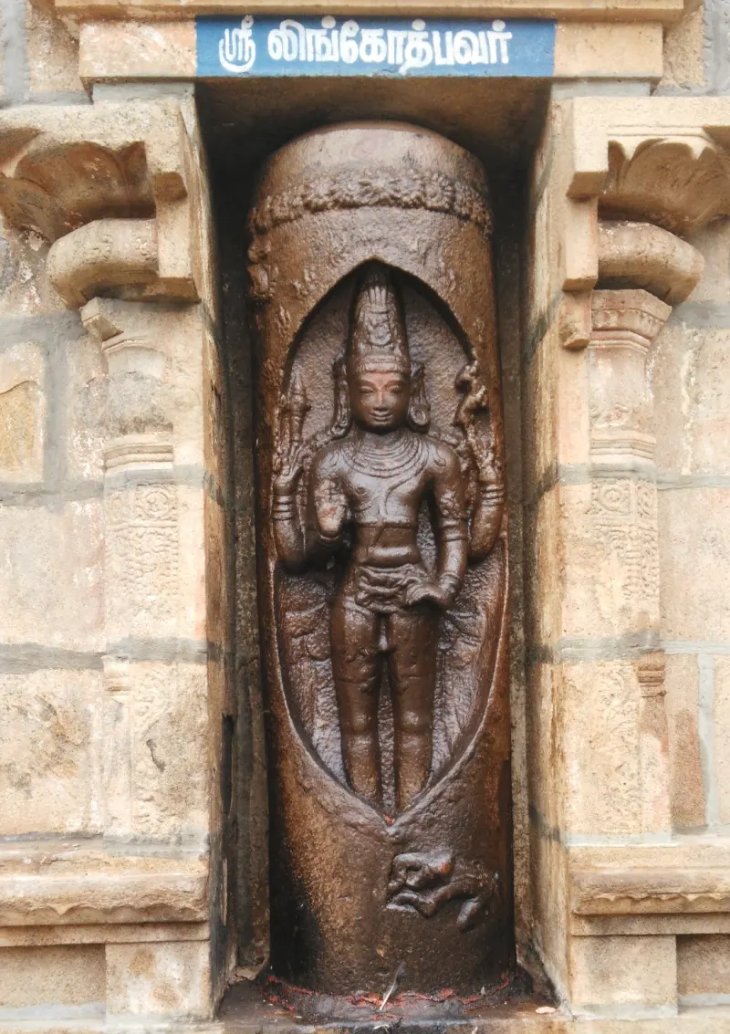 Carved figure standing inside an oval opening of a tall stone pillar, under a Tamil name board