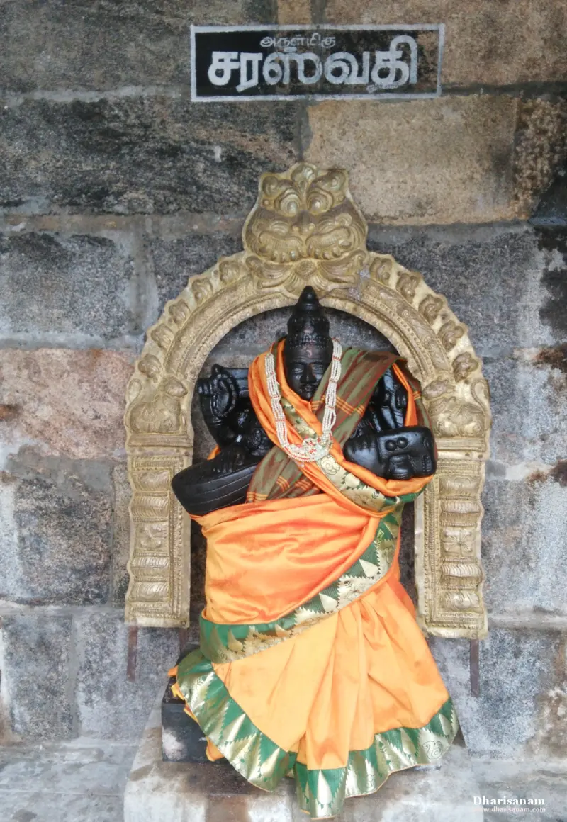 A black stone idol of Saraswathi in an orange silk sari, under a gilded arch and a Tamil name board.