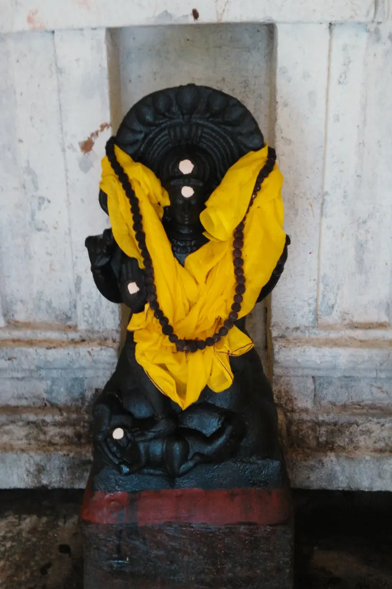 A dark stone seated figure wearing a yellow cloth and a rudraksha garland