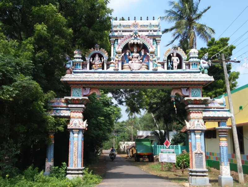 A tall painted arch with deity figures spans a village road, with a palm tree and trees behind.