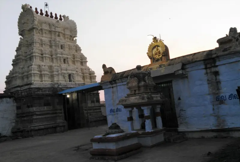 A tiered gopuram on the left and a blue temple wall with a small stone shrine in front, at dusk.