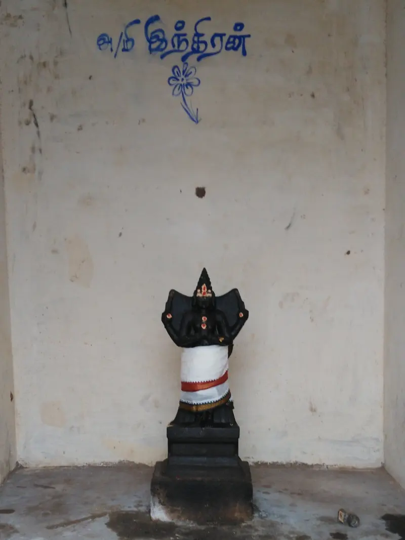 A small black idol in white cloth stands before a wall with Indran written in Tamil
