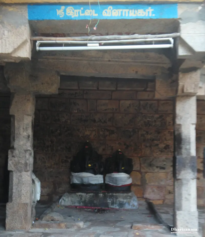Two black stone Vinayakar idols side by side on a low platform, under a blue Tamil name board