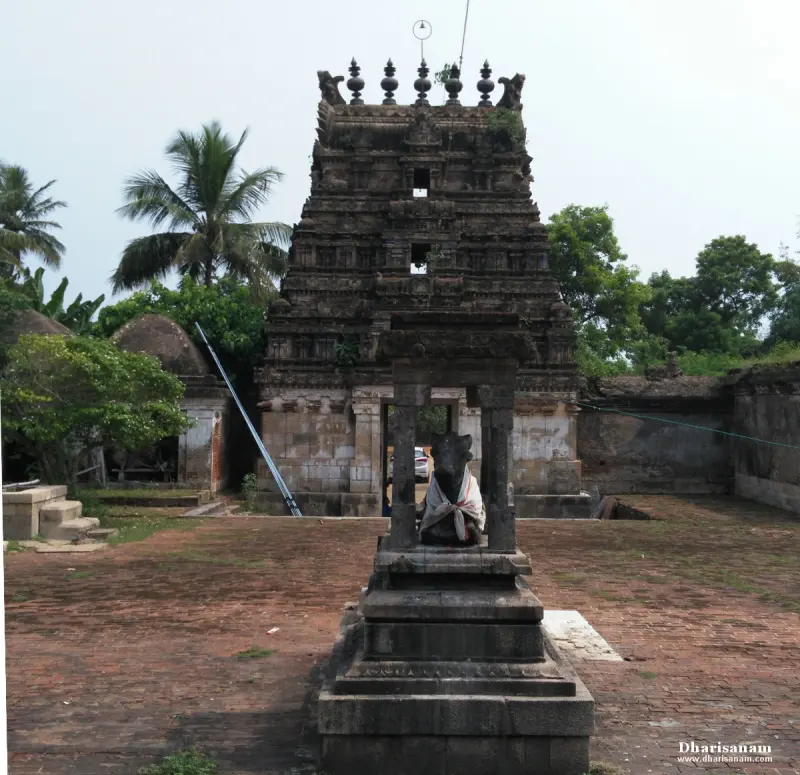 A dark, weathered gopuram with weeds on its tiers; a Nandi sits on a stone pedestal under a small pavilion in front.
